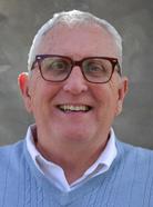 A man with gray hair and glasses, wearing a light blue sweater over a white collared shirt, smiles at the camera against a neutral background.