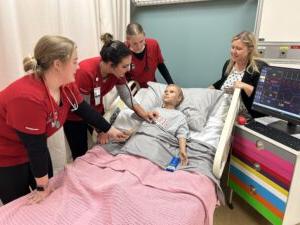 Four nursing students in red uniforms practice on a medical mannequin in a hospital bed, with a heart rate monitor nearby. An instructor observes the session.